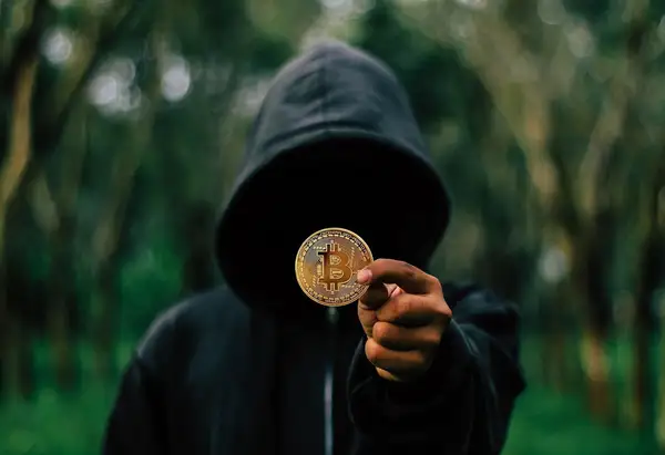 شخص بقلنسوة يحمل عملة بيتكوين في الظلام، يمثّل المحتالين المجهولين الذين يستهدفون مشتري العملات الرقمية في السعودية